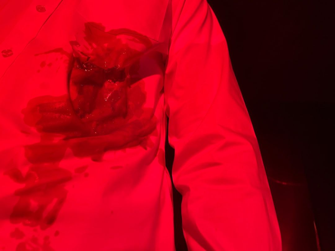 En närbild av en vit skjorta med en stor röd fläck och mörka detaljer, upplyst av rött ljus.